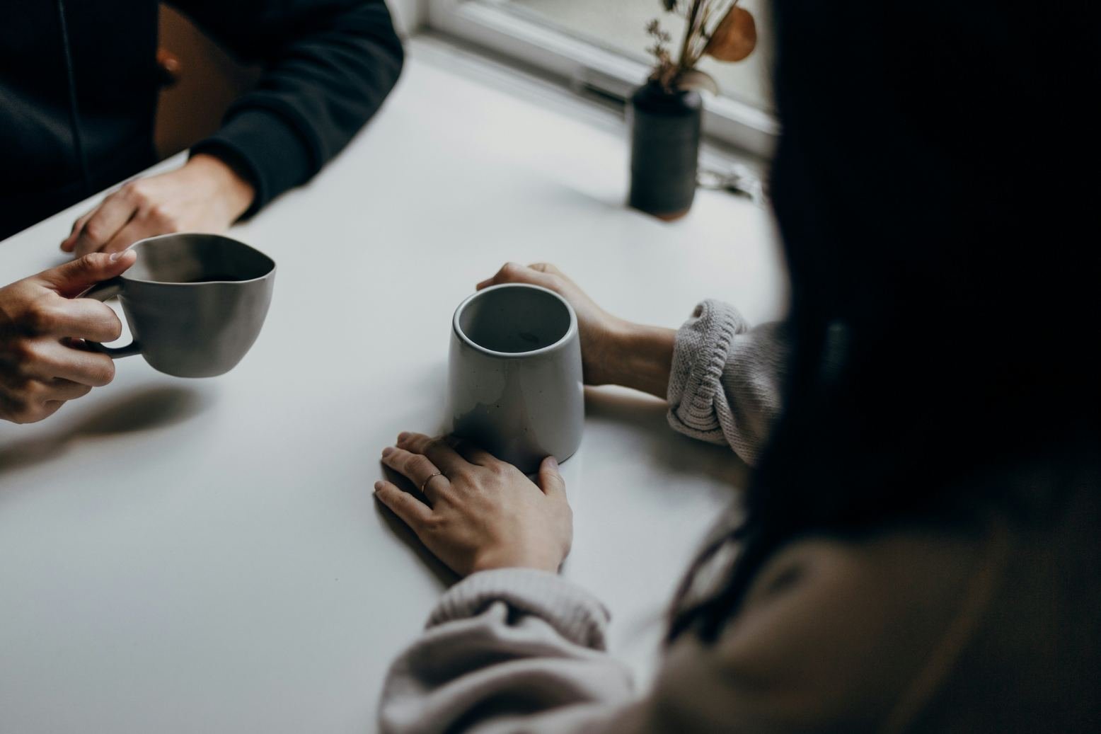 Dos personas conversan alrededor de una mesa blanca mientras sostienen tazas; la escena está iluminada por una ventana y contiene una pequeña planta al fondo